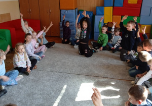Uczniowie siedzą na dywanie i podnoszą ręce
